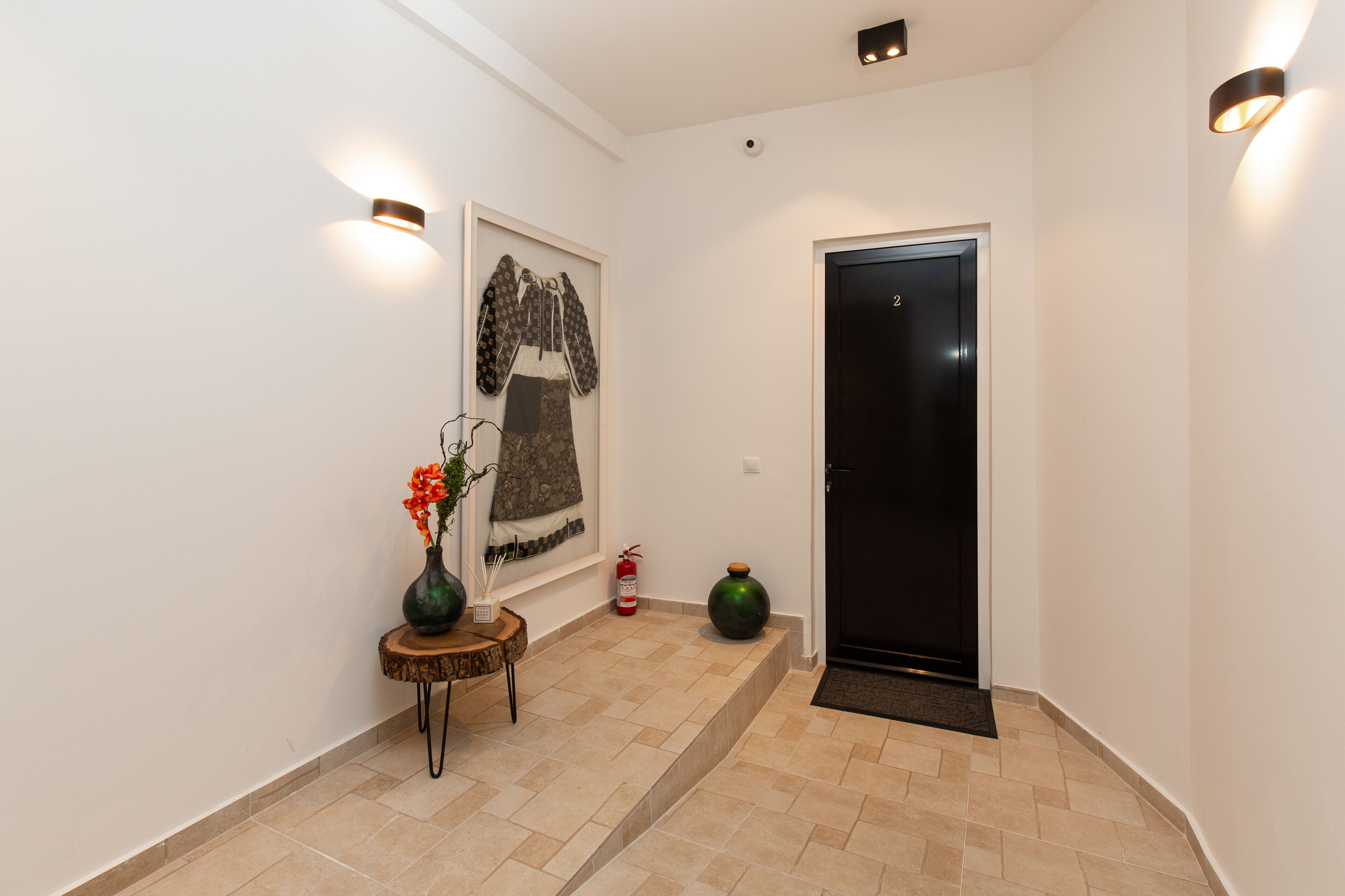 A hallway with a framed artwork, a small table with a vase, a fire extinguisher, and a door.