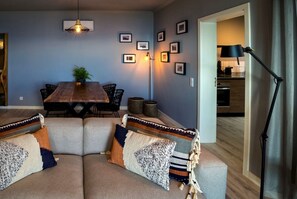 A cozy living room with a sectional sofa, a wooden dining table, and a kitchen in the background.