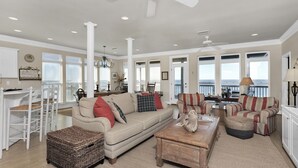 A spacious living room with a large window overlooking the ocean, a ceiling fan, and a dining area with a view. - Corner pet-friendly, 5/3 bay sunset views! - TraMarLis (Dauphin Island)