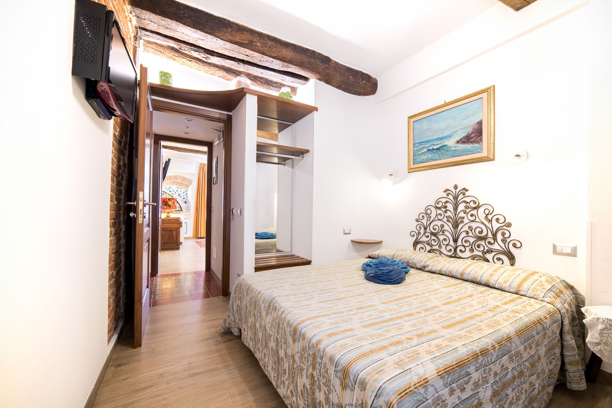 A bedroom with a bed, a wooden headboard, a painting, and a view of a room with a window.