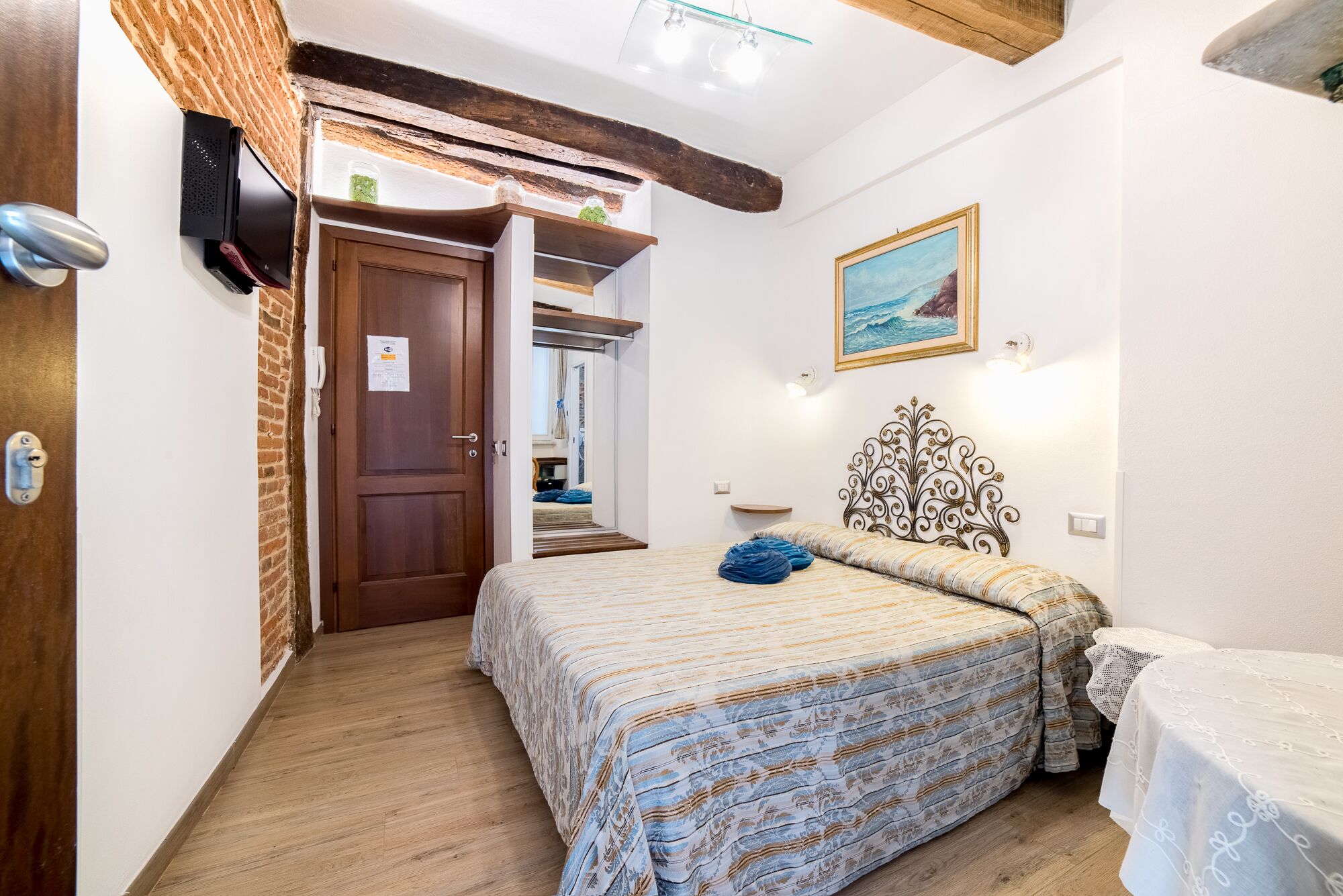 Un dormitorio con puerta de madera, cama con ropa de cama estampada, cabecero decorativo, un cuadro en la pared y una mesita con mantel blanco.
