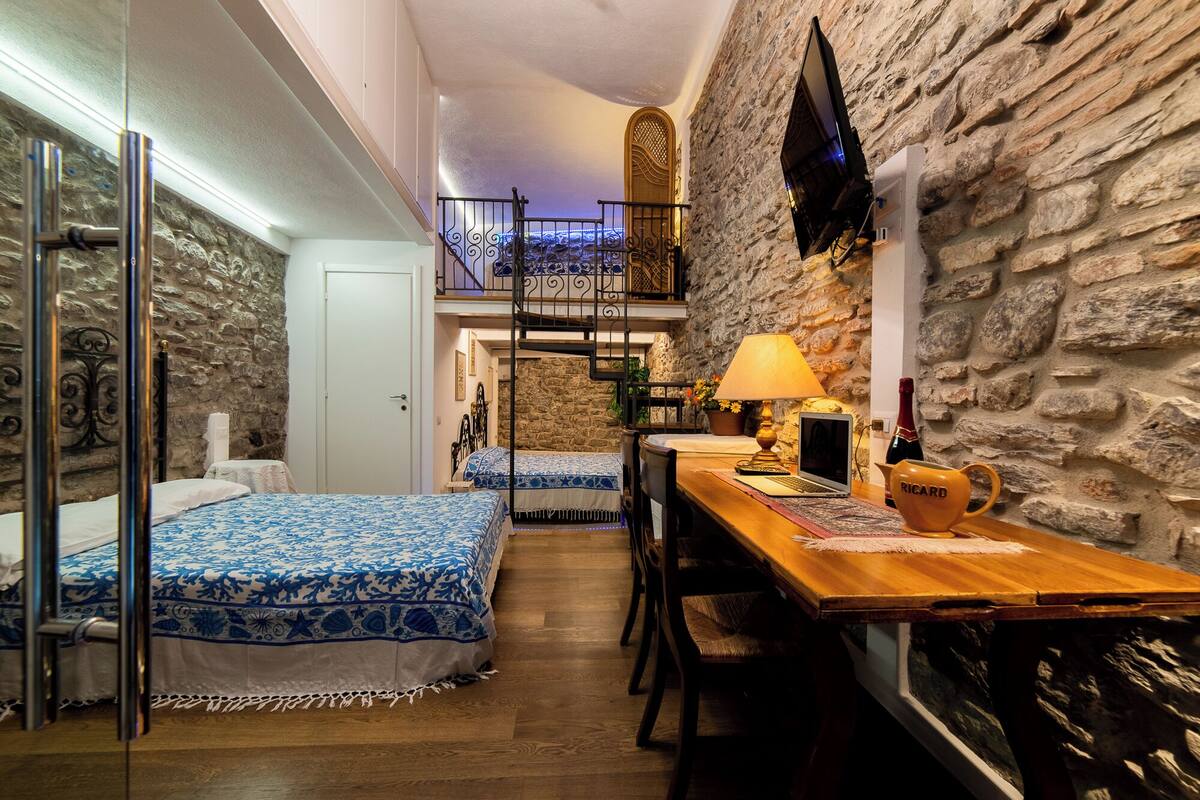 A cozy room with two beds, a wooden table, a lamp, a TV, and a stone wall.