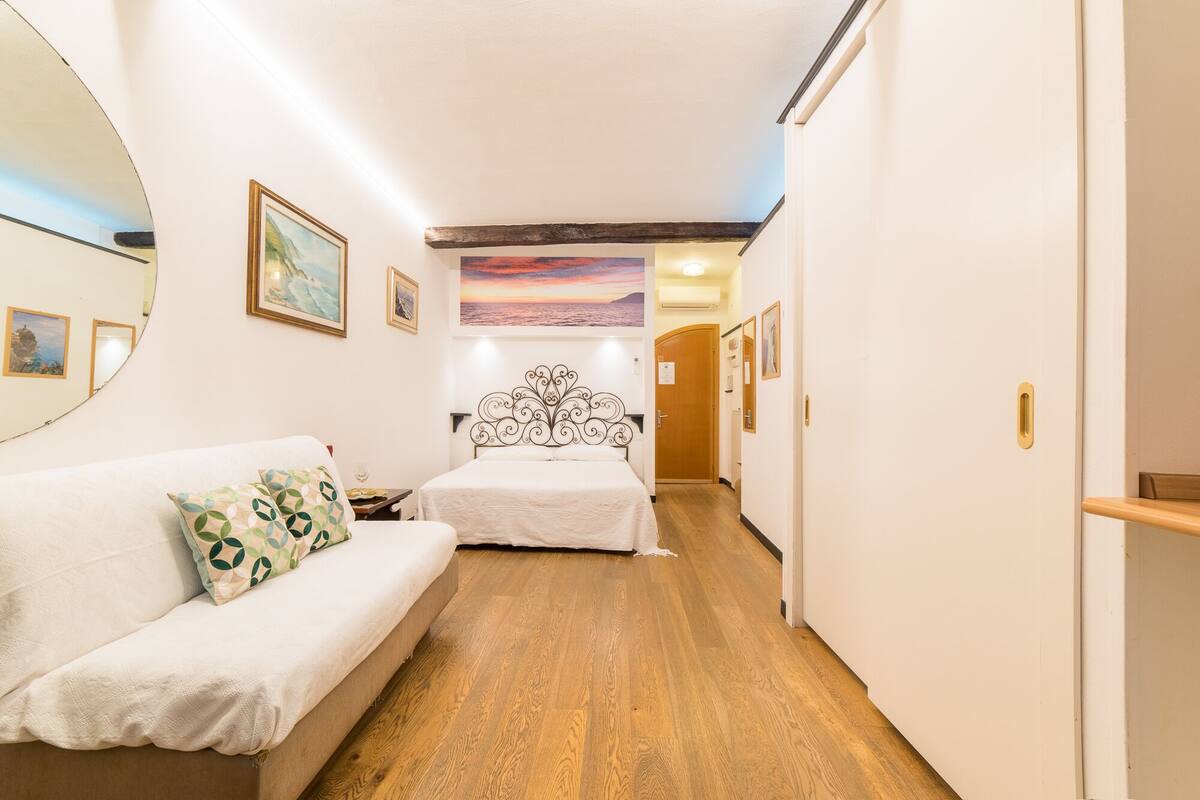 A hotel room with a wooden floor, a white bed, a white sofa, and a painting of a sunset.