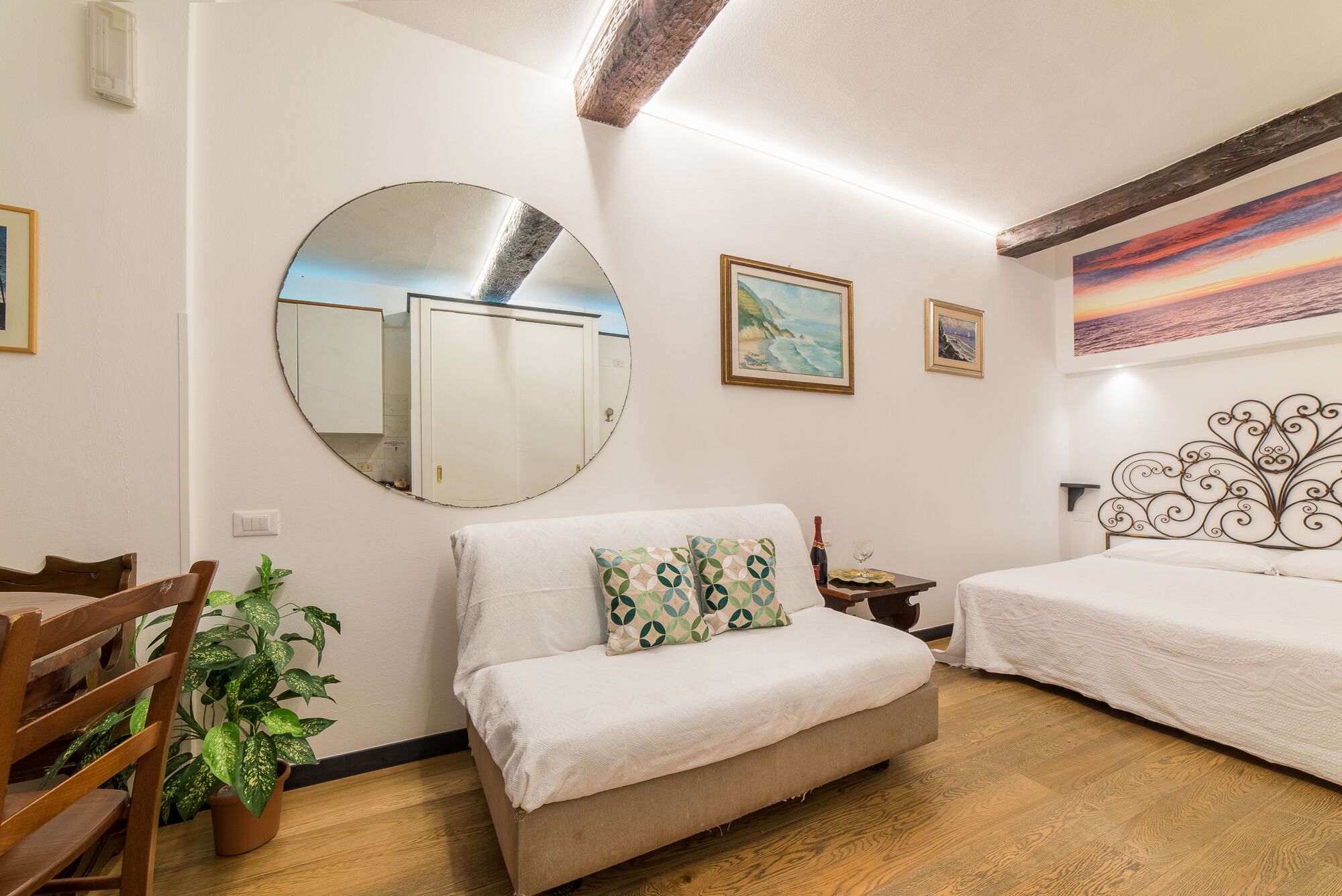 A bedroom with a round mirror, two beds, a wooden floor, and a painting of a sunset.