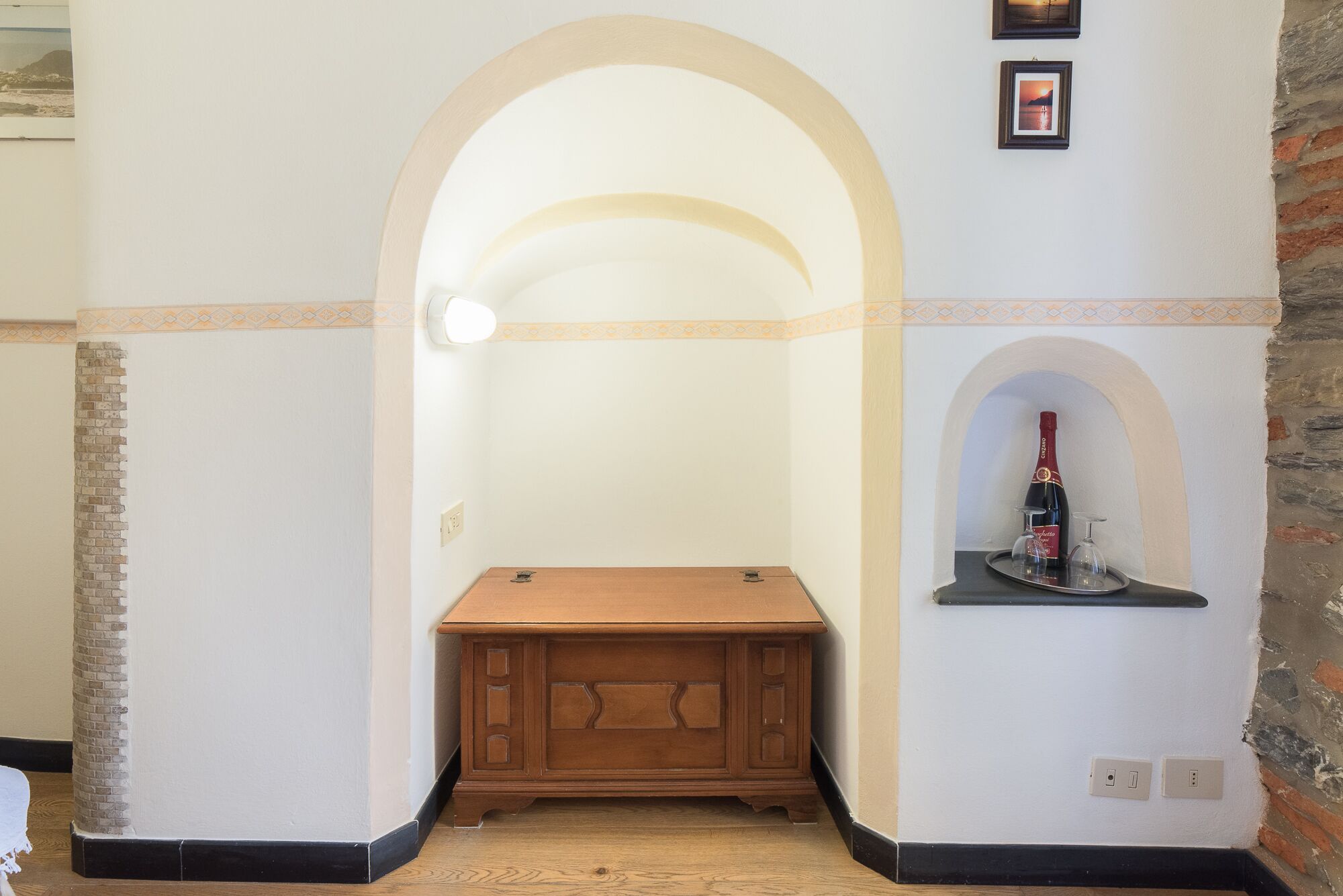 Una habitación de esquina con un armario de madera, una pequeña estantería con una botella y vasos, y un arco de entrada.