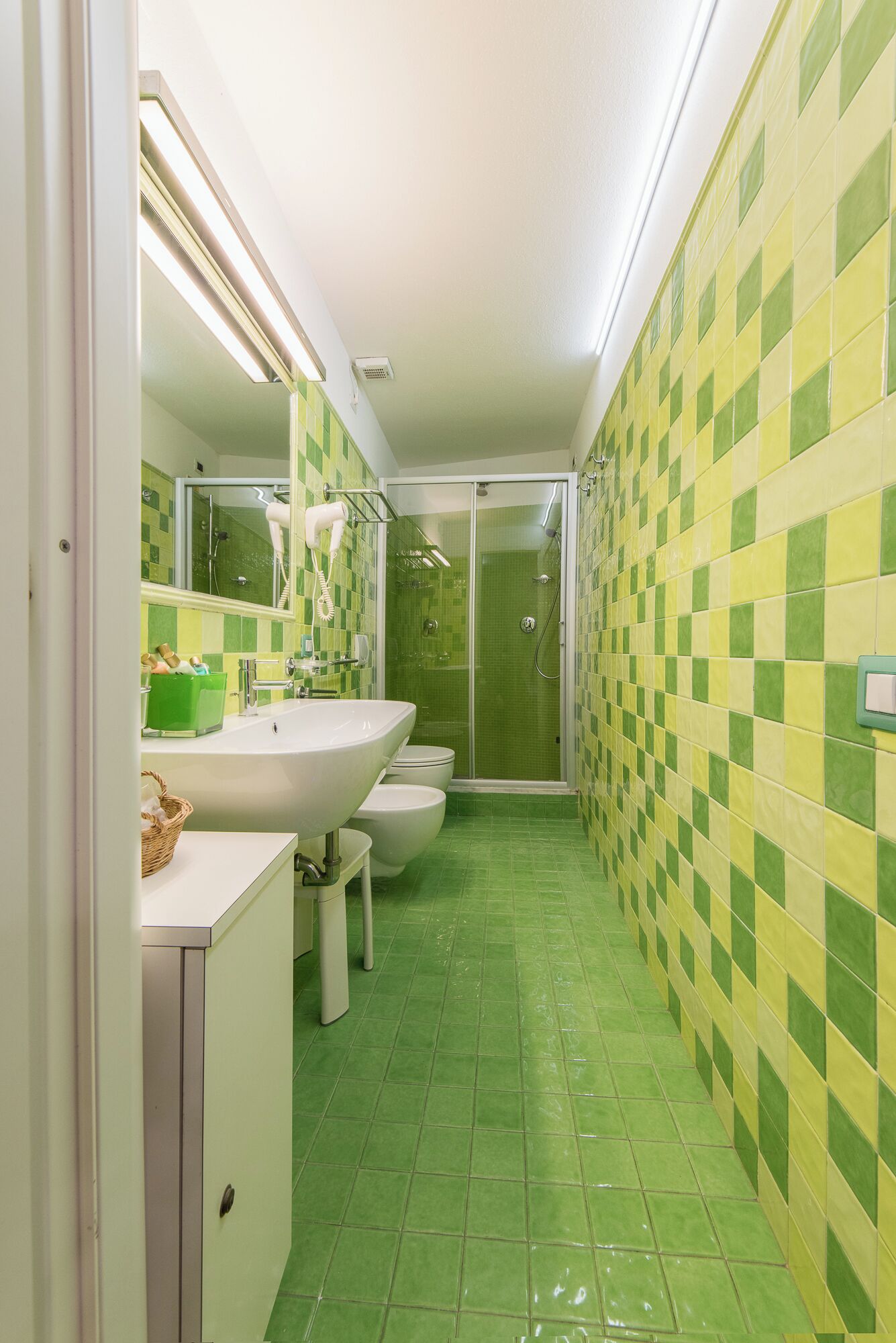Ein schmales Badezimmer mit Waschbecken, Toilette und Dusche. Die Wände sind mit einem Schachbrettmuster gefliest.