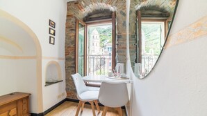 A small room with a round mirror, a white chair, a wooden cabinet, and a window with a view of buildings and greenery. - Vernazza Sea View (Vernazza)
