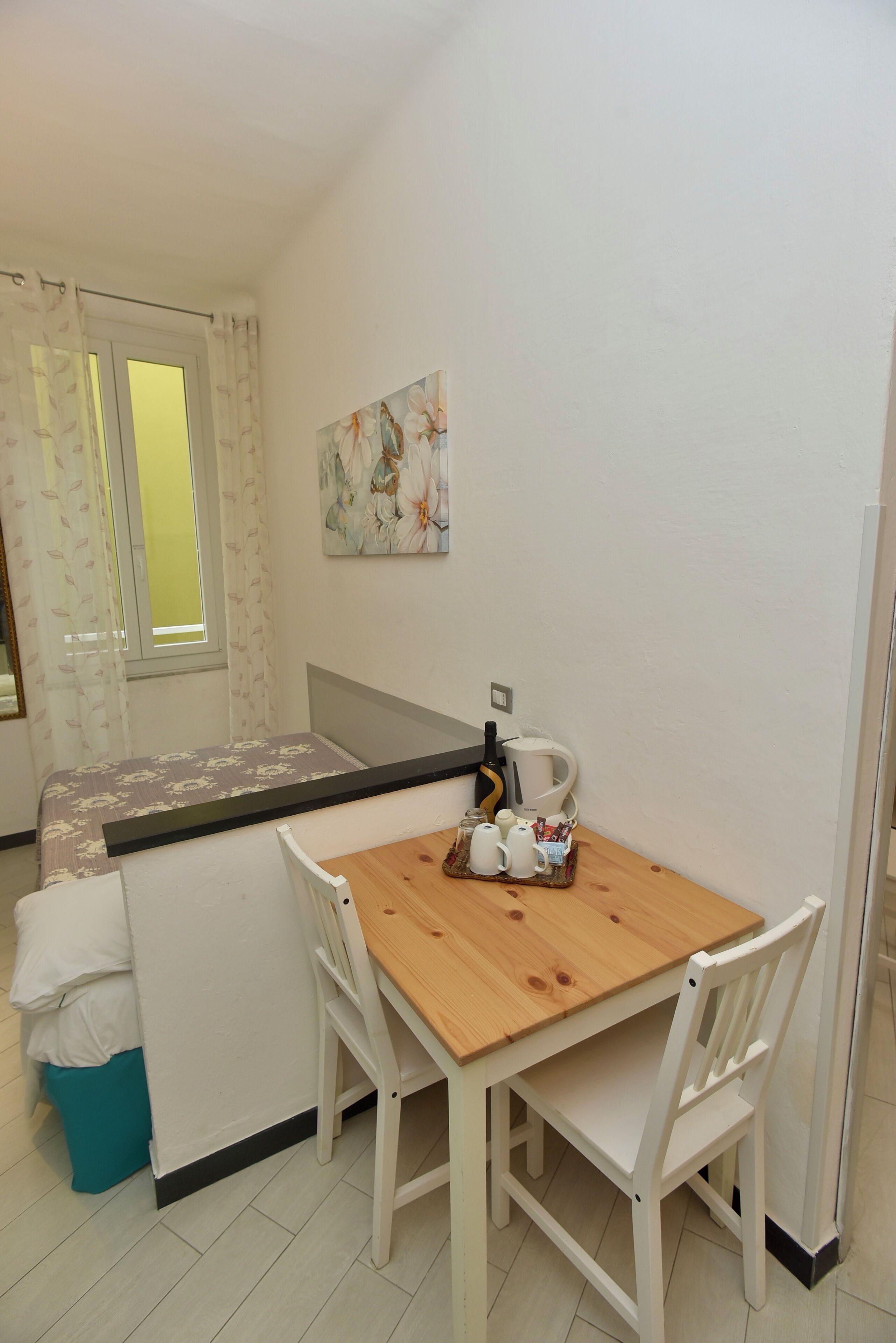 A small, clean room with a bed, a table with chairs, a window with curtains, and a painting on the wall.