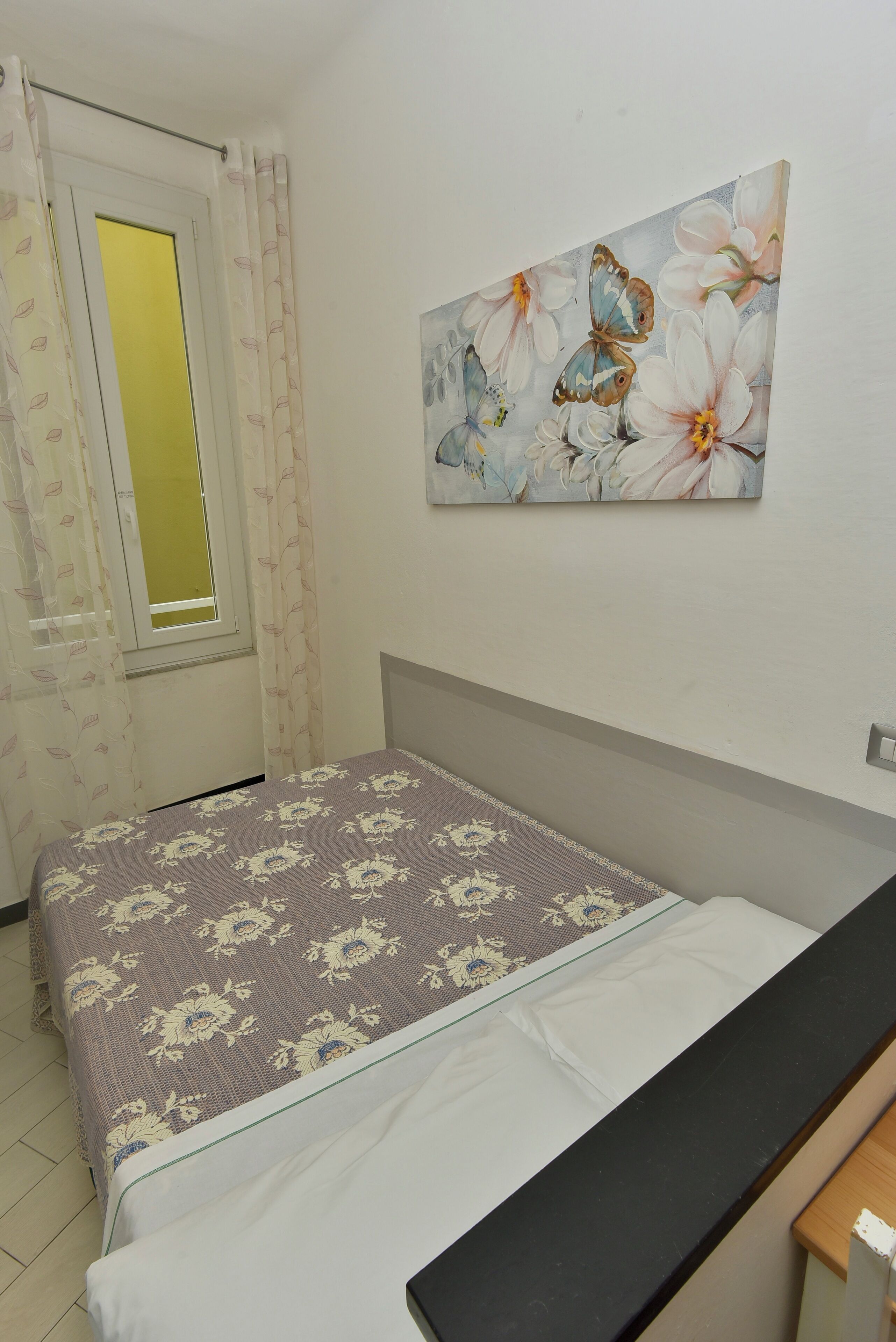 Un dormitorio con cama, mesita de noche, ventana con cortinas y un cuadro colgado en la pared.