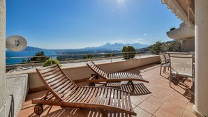 Une terrasse dotée de chaises longues en bois, d’une table et de chaises, offrant une vue panoramique sur un plan d’eau et des montagnes.