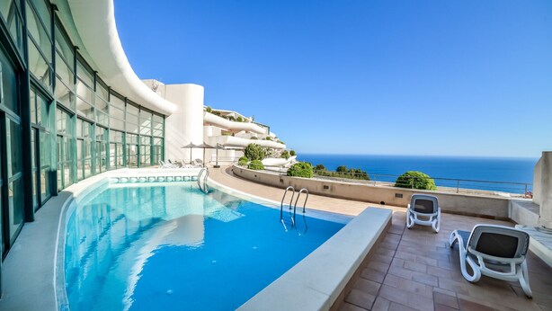 Une piscine à l’eau d’un bleu cristallin, entourée d’un bâtiment aux lignes courbes et doté de grandes fenêtres, offrant une vue sur l’océan.