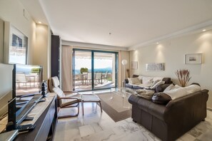 Un salon moderne avec un canapé, une table basse et un téléviseur. Il y a un balcon avec des meubles d’extérieur et une vue sur la mer.