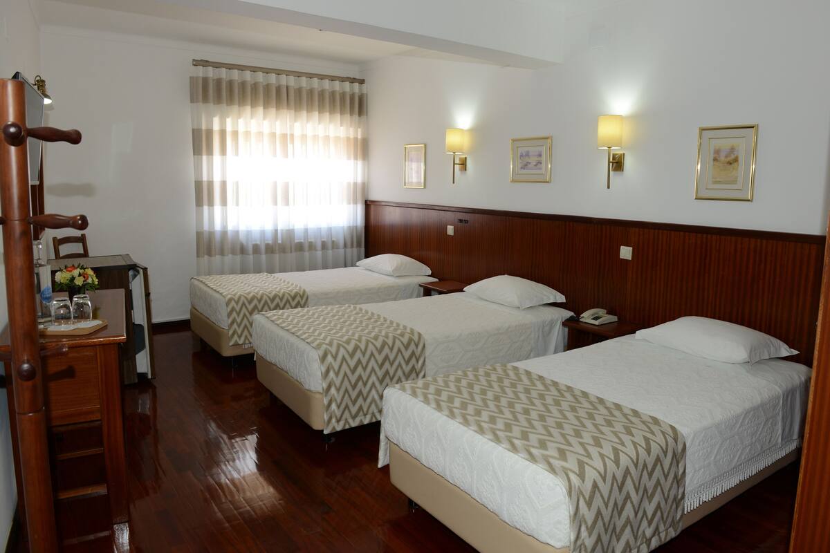 A hotel room with two beds, wooden headboard, bedside tables, a desk with a chair, a lamp, and a window with curtains.