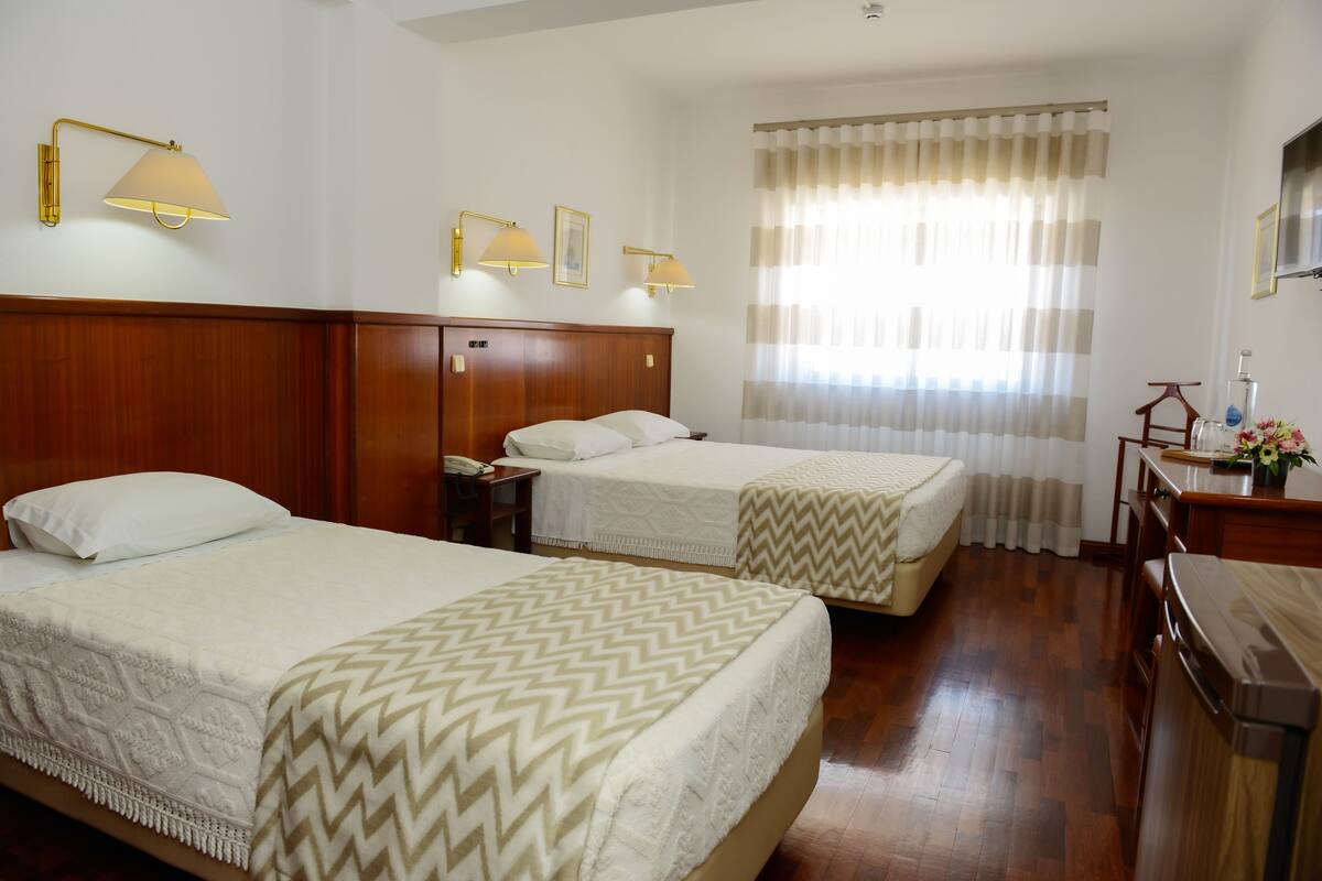 A hotel room with two beds, wooden headboards, a wooden dresser, a nightstand, and a window with curtains.