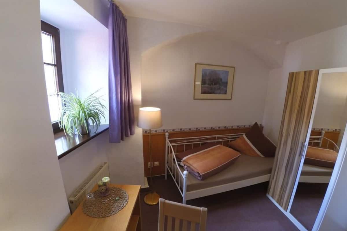 Una habitación pequeña y acogedora con una cama individual, un escritorio, una silla, una lámpara y una ventana con cortinas.
