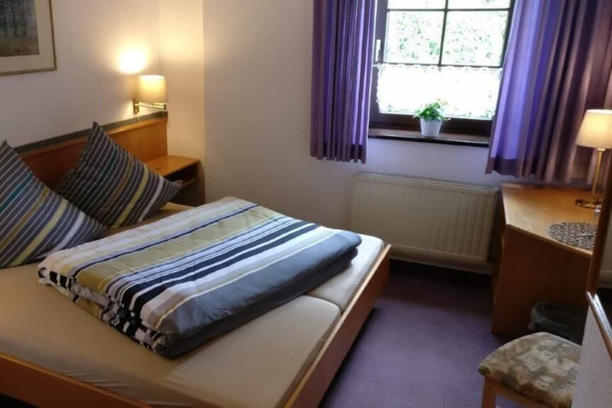 Una cama bien hecha con ropa de cama a rayas, una mesita de noche con una lámpara, una ventana con cortinas moradas y una pequeña maceta en el alféizar.