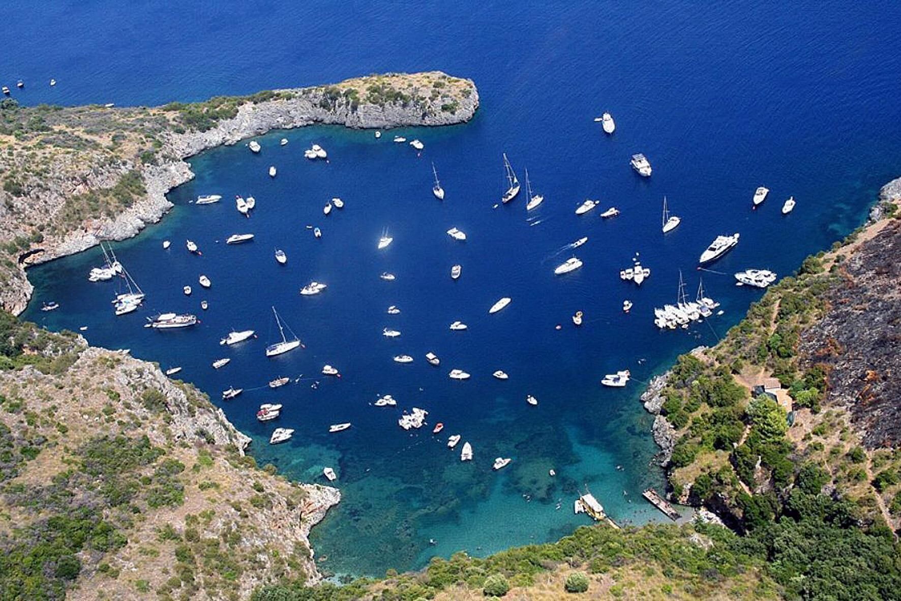 Une zone côtière où de nombreux bateaux sont amarrés dans une baie, elle-même entourée d’un paysage rocheux et d’une végétation clairsemée.