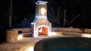 A stone-built outdoor fireplace with a fire burning inside, surrounded by a stone patio and a swimming pool. - Club Vergel Resort (Ezequiel Montes)