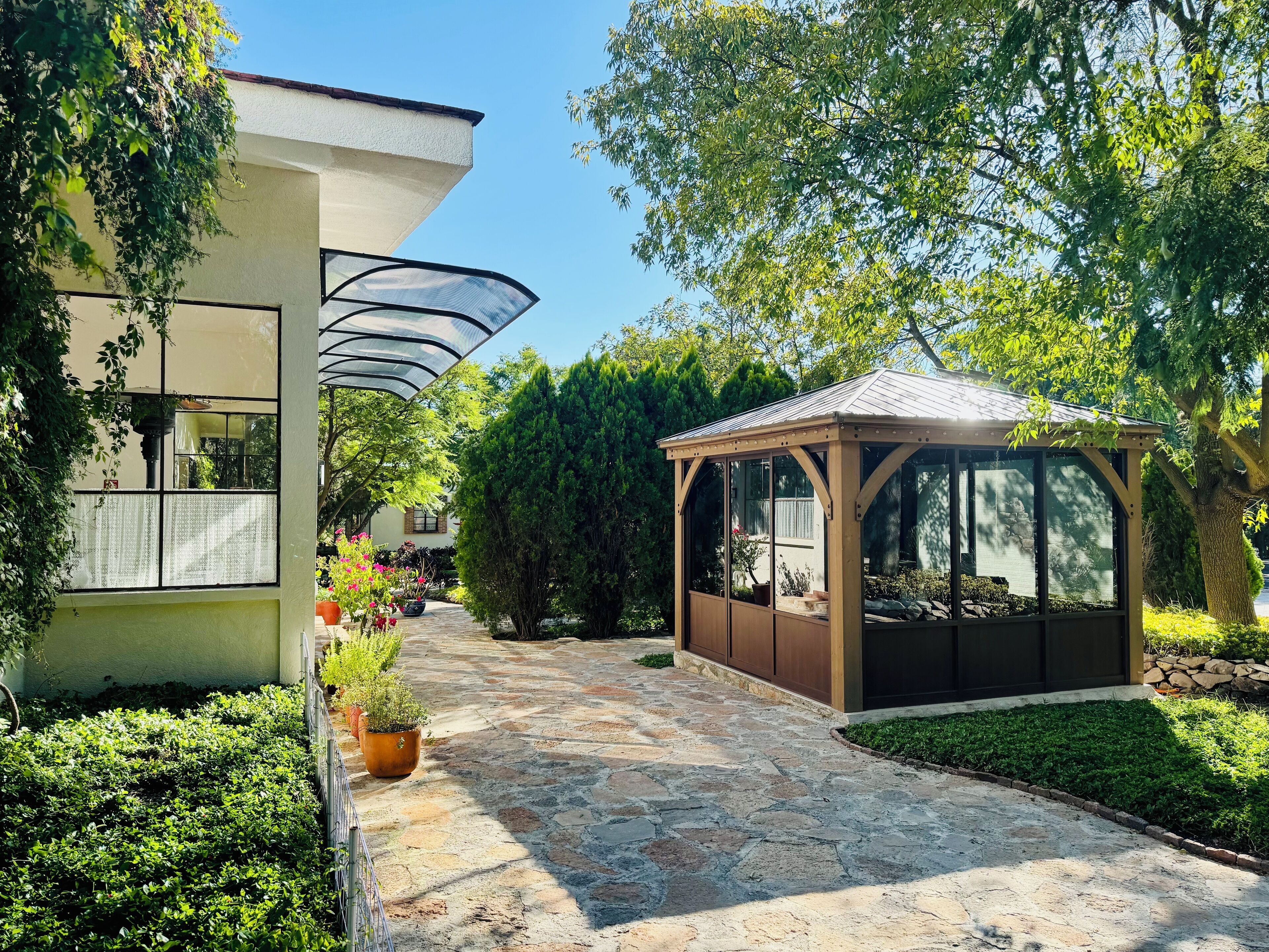 A covered outdoor area with a stone pathway, a wooden gazebo, and a building with a glass enclosure.