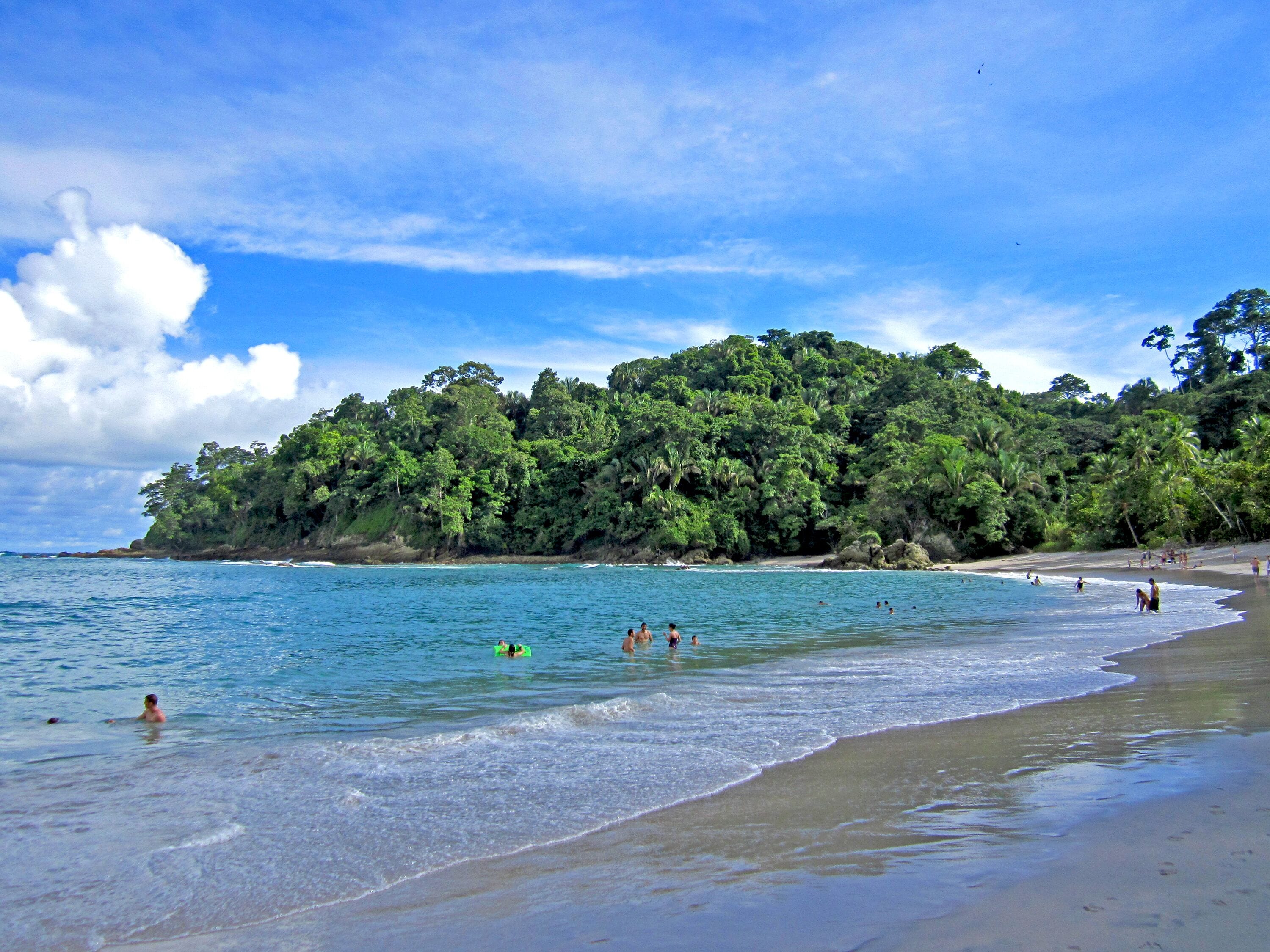 A beach with clear blue water, people swimming and relaxing, and a lush green hill in the background.