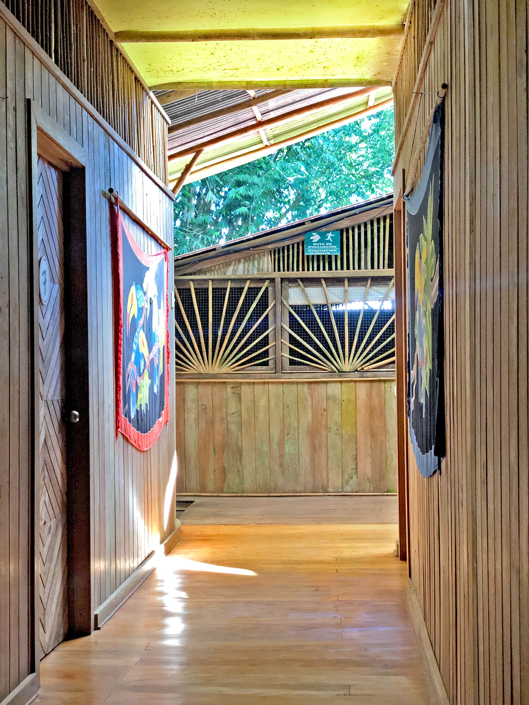 A hallway with wooden walls and a wooden floor, leading to a covered area with a view of trees.