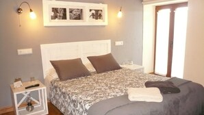 Un dormitorio con cama, dos almohadas, una mesita de noche con lámpara, un cuadro enmarcado en la pared y una ventana con cortinas.
