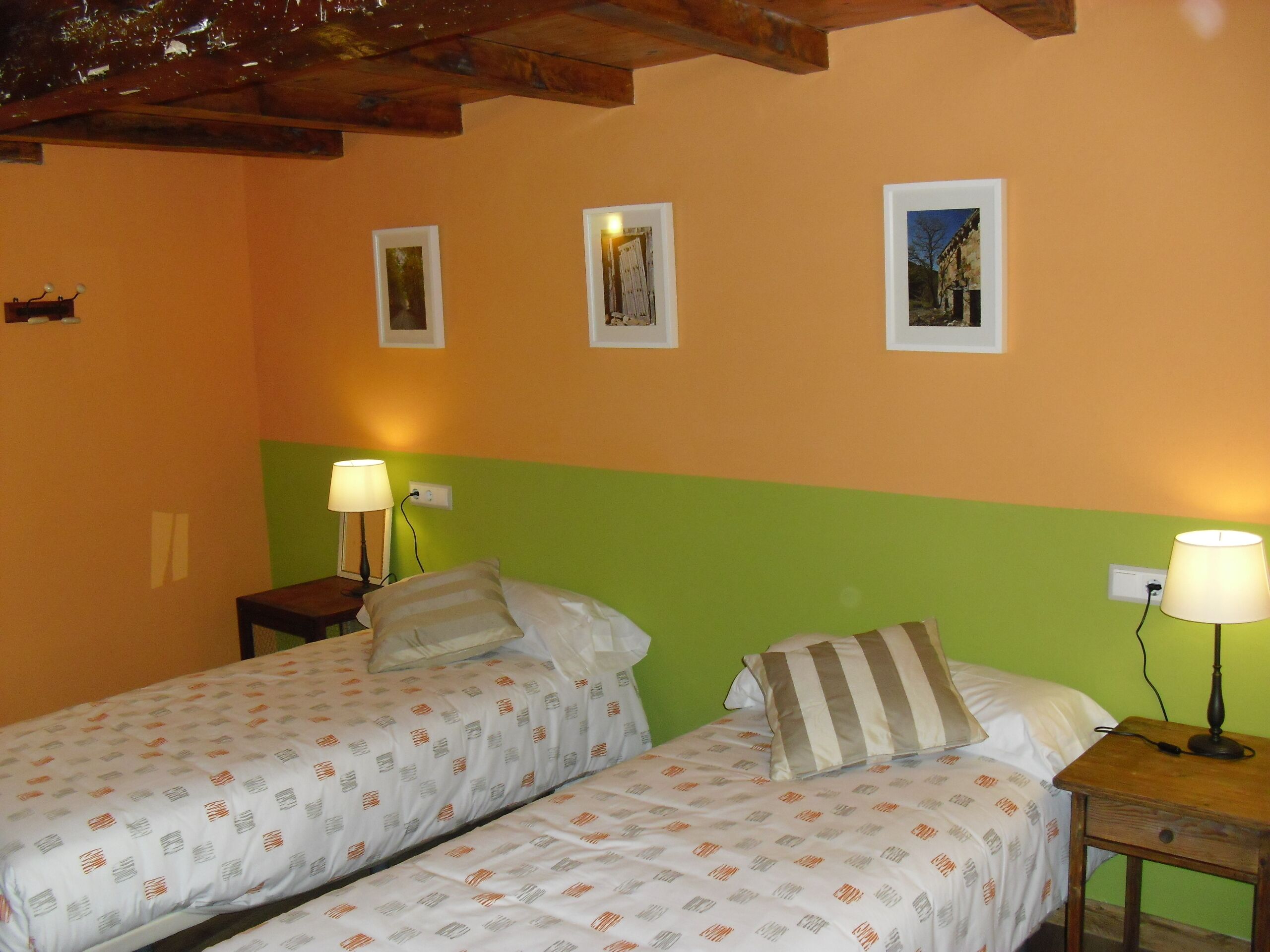 Una habitación con dos camas, cada una con una almohada y una mesita de noche con lámpara. Las paredes están pintadas en dos colores: naranja y verde.