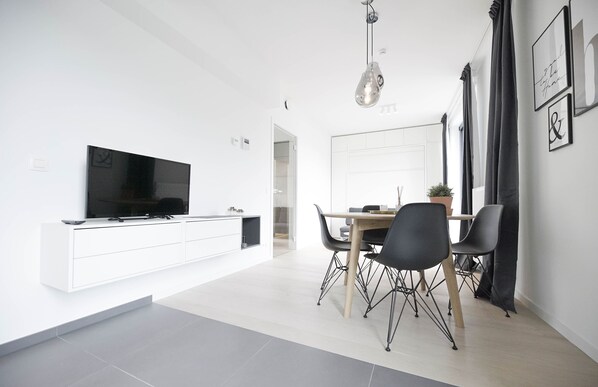 A modern living room with a flat-screen TV on a white console, a dining area with a wooden table and black chairs, and a pendant light.