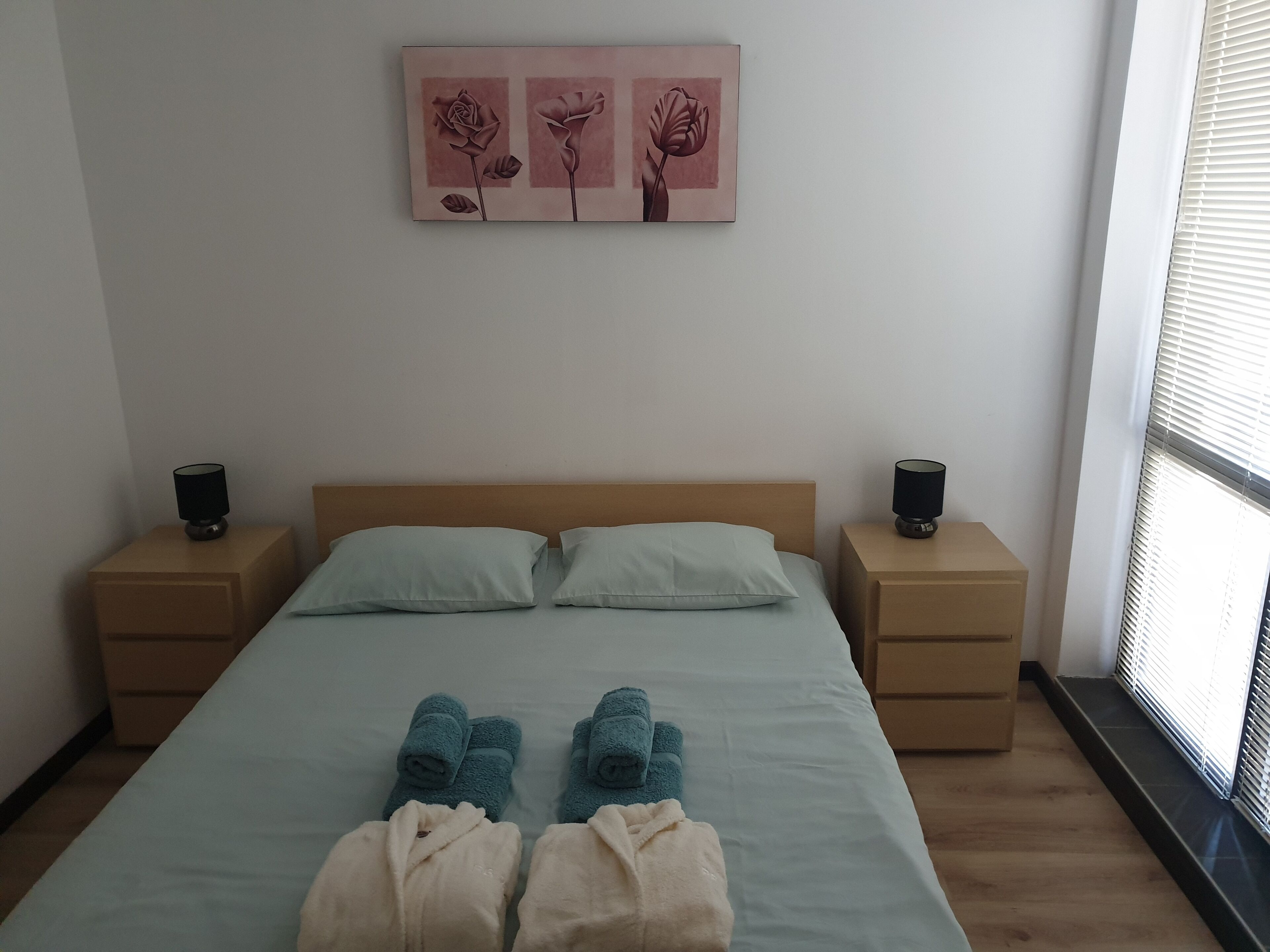 A bedroom with a bed, two bedside tables, a window with blinds, and a wall-mounted artwork of flowers.