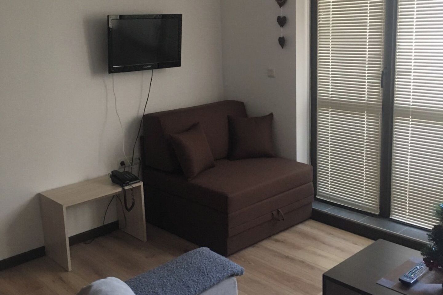 Una sala de estar con un sofá marrón, una mesita, un televisor montado en la pared y una ventana con persianas.
