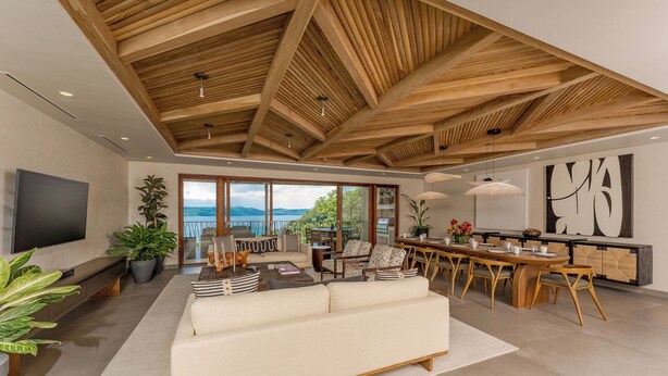 Amplia sala de estar con un sofá grande, una mesa de comedor y sillas, vigas de techo de madera y un ventanal con vistas al exterior.