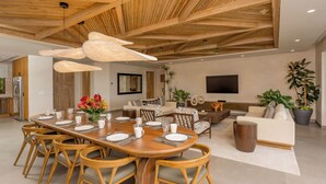 Un comedor moderno con techo de madera, una larga mesa de comedor preparada para seis personas y una zona de estar cómoda con sofás y televisor.