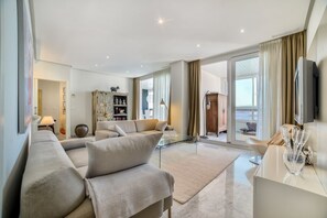 A spacious living room with a sofa set, a glass coffee table, and a view of the outdoors through large windows.