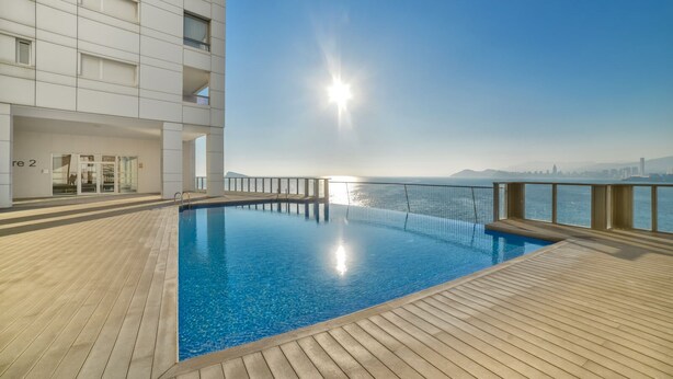 En moderne swimmingpool med træterrasse, glasindgang og udsigt til havet og byens skyline.