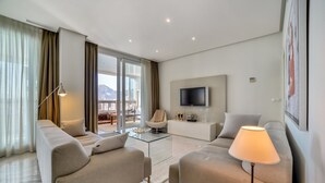 En moderne stue med sofaarrangement, sofabord, fjernsyn ophængt på væggen og en balkon med udsigt over bjergene.