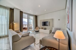 A modern living room with a sofa set, a coffee table, a TV mounted on the wall, and a balcony with a view of the mountains.