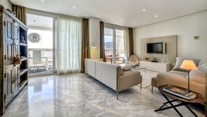 En moderne stue med sofa, lænestole, sofabord og fjernsyn. Der er en balkon med udsigt over byen.