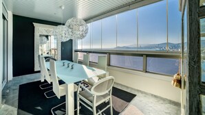 Et moderne spiseområde med et stort bord og stole, en dekorativ lysekrone og udsigt over bybilledet gennem store vinduer.