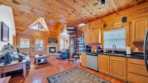 A cozy cabin interior with a fireplace, wooden floors, a dining area, and a kitchen.
