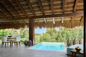 A pool area with a thatched roof, a dining table with chairs, and a view of lush greenery.
