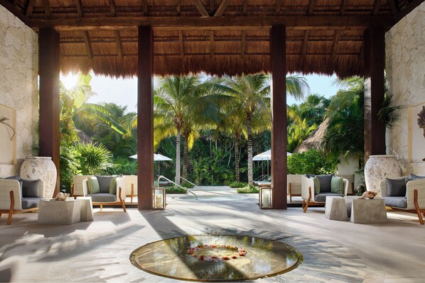 A tropical resort lobby with a thatched roof, comfortable seating, a central water feature, and a view of palm trees.