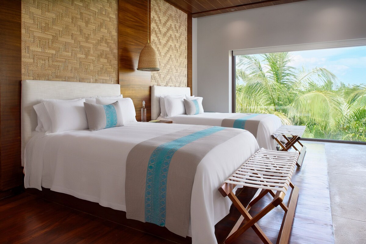 Habitación de hotel con dos camas, un ventanal con vistas a palmeras y un panel decorativo en la pared tejido.