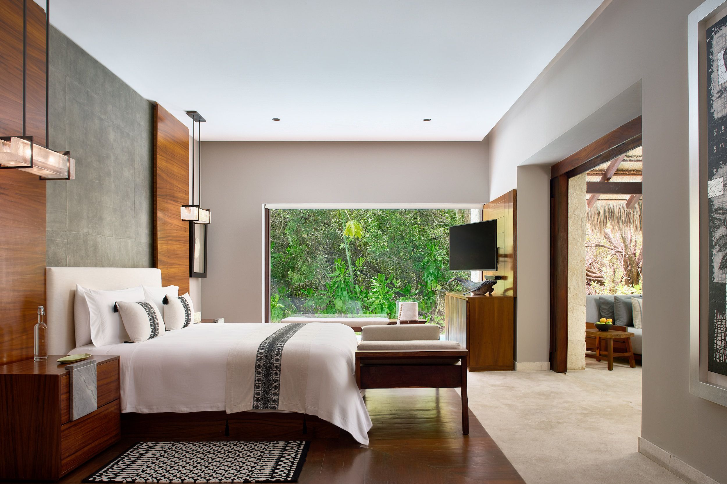 A modern bedroom with a large bed, bedside tables, a TV, and a view of greenery.