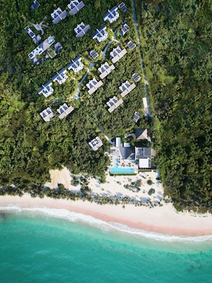 Vista aérea de un resort con múltiples alojamientos tipo bungalow rodeados de densa vegetación tropical, una playa de arena y aguas turquesas cristalinas.