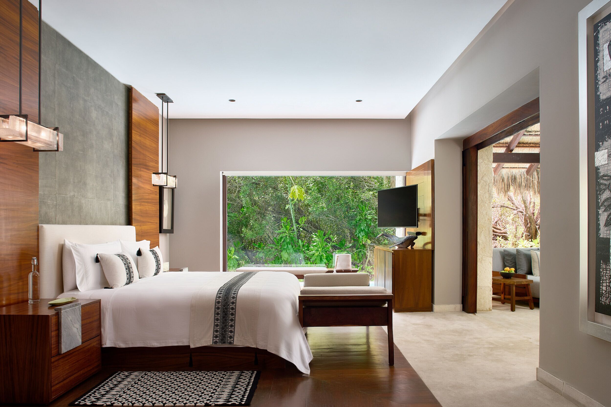 A modern bedroom with a large bed, bedside tables, a TV, and a view of greenery.