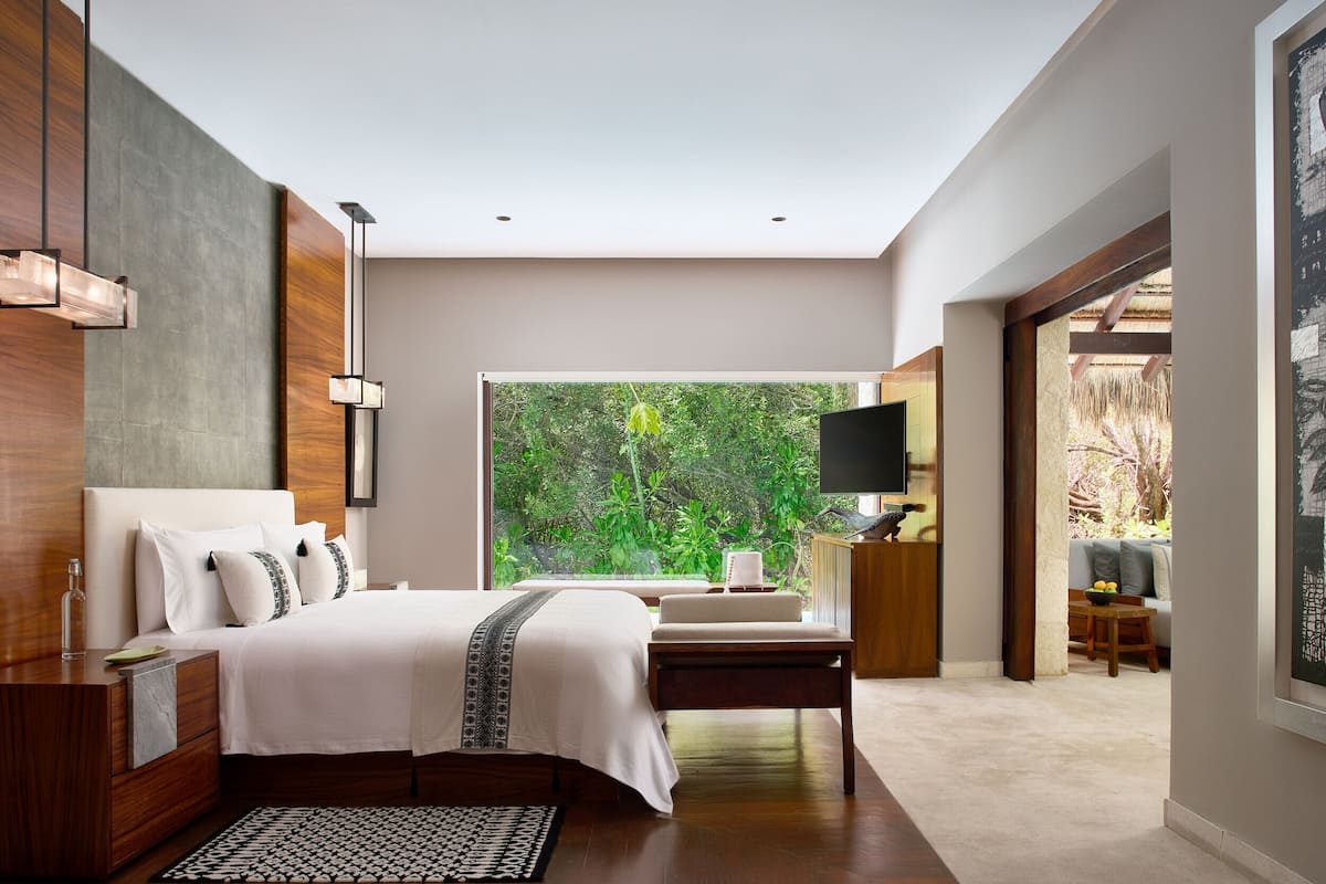 A modern bedroom with a large bed, bedside tables, a TV, and a view of greenery.