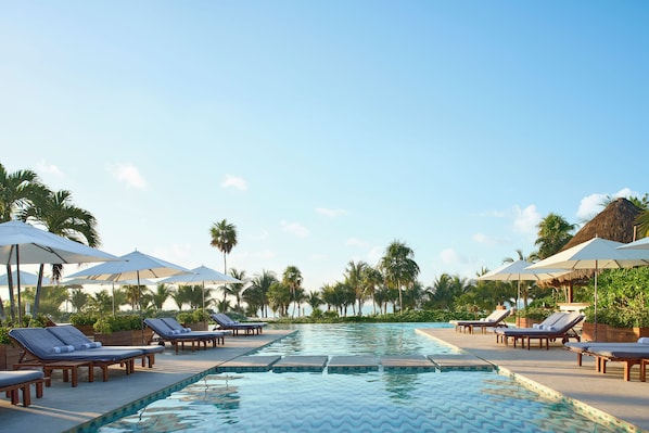 Un área de piscina con sillones reclinables y sombrillas, rodeada de palmeras.
