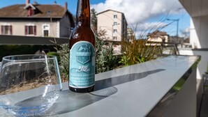 A bottle of beer and a glass on a balcony with a view of buildings and a clear sky.