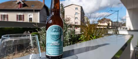 A bottle of beer and a glass on a balcony with a view of buildings and a clear sky.