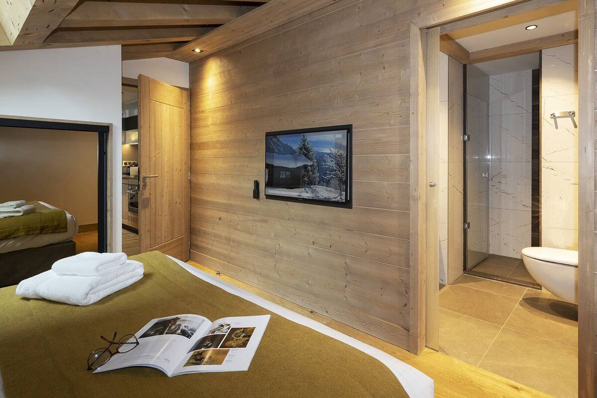 A hotel room with a wooden wall, a framed picture, a bed with towels, a magazine, and a bathroom with a toilet and shower.